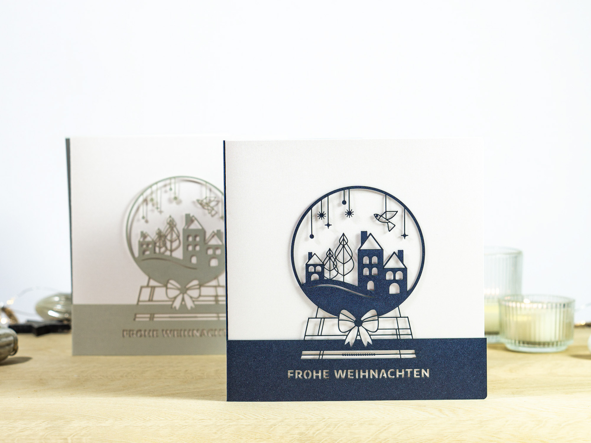 Zwei Farbvarianten PALUXA Lasercut-Weihnachtskarte mit Schneekugel Motiv, stehend, in Farbe Nachtblau und Silber.